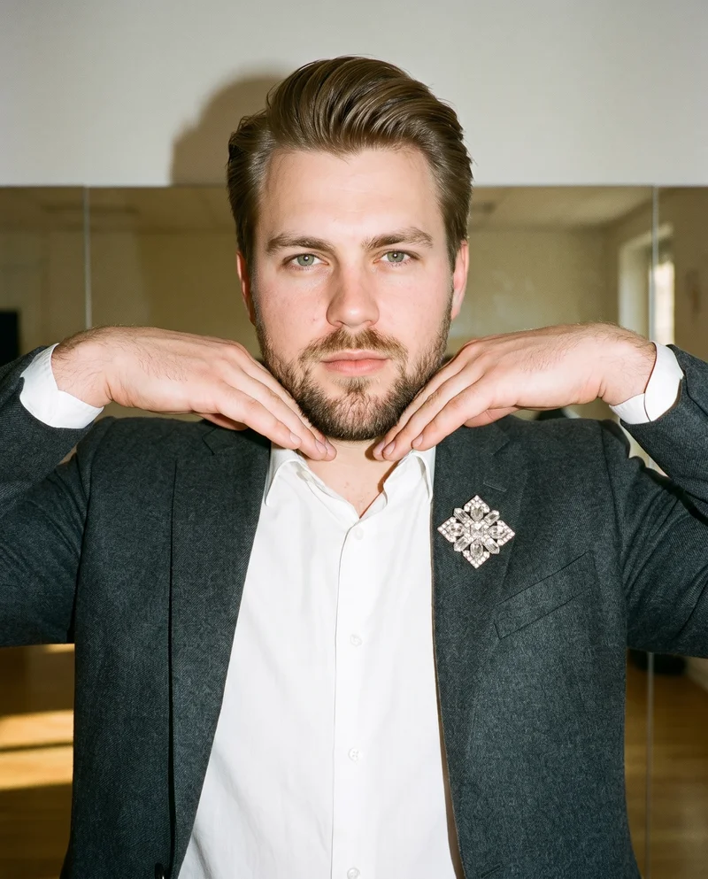 A 29-year-old Caucasian man is shown in a front-view bust composition wearing a men's crystal brooch. His hands are positioned at his jaw, and he has a neutral expression. A yoga studio serves as the backdrop.