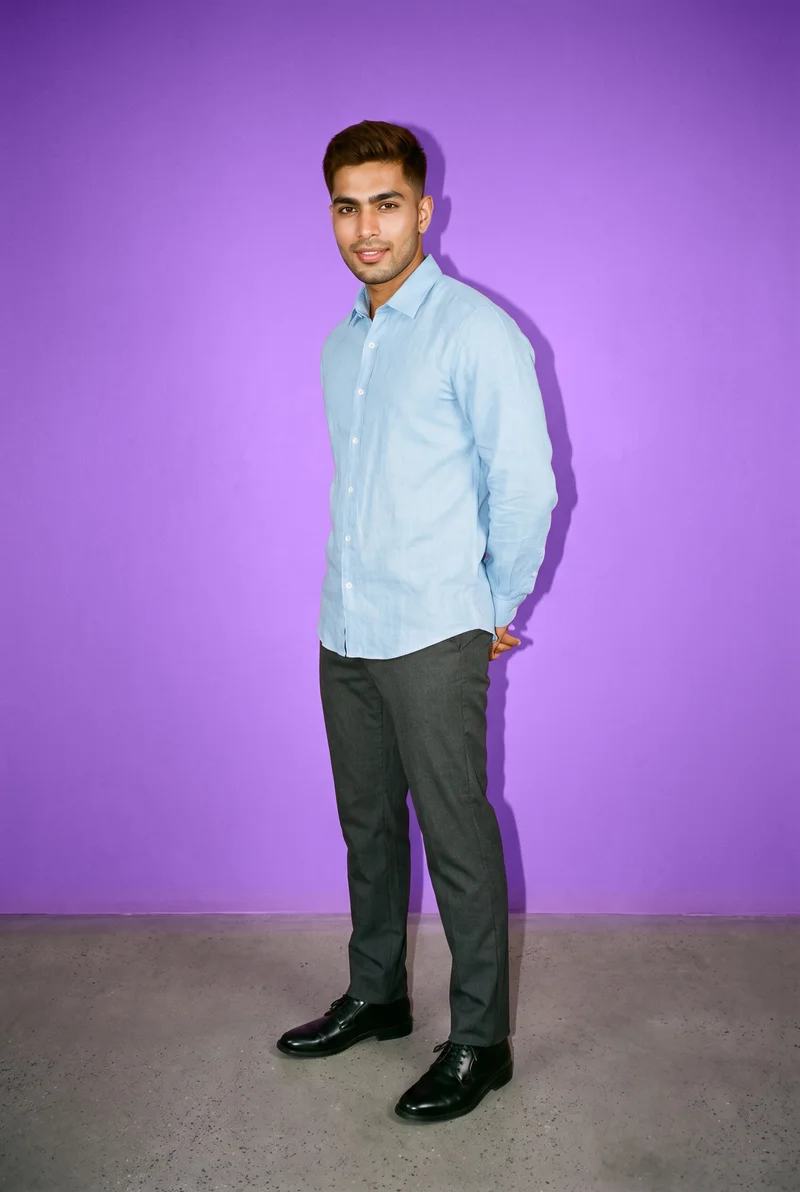 A 29-year-old Indian man wears a sky blue linen shirt in a full-body view from a front three-quarter angle, standing with his hands behind his back and a natural facial expression. An amethyst studio-wall backdrop fills the background.