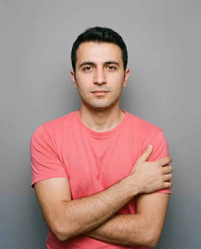 A 36-year-old Turkish man wears a coral seamless performance top in an upper-body front view with his arms crossed and a confident expression. A pebble grey studio-wall backdrop frames the composition.
