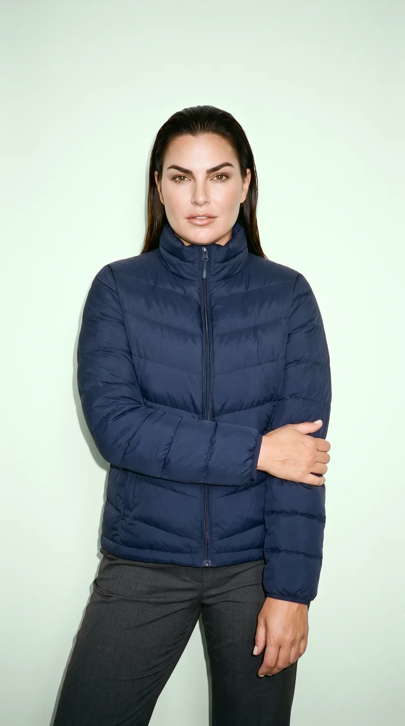 A 50-year-old Caucasian woman wears a navy insulated down jacket in a front-facing three-quarter-length view. She holds one arm with a neutral expression against a honeydew studio-wall backdrop.