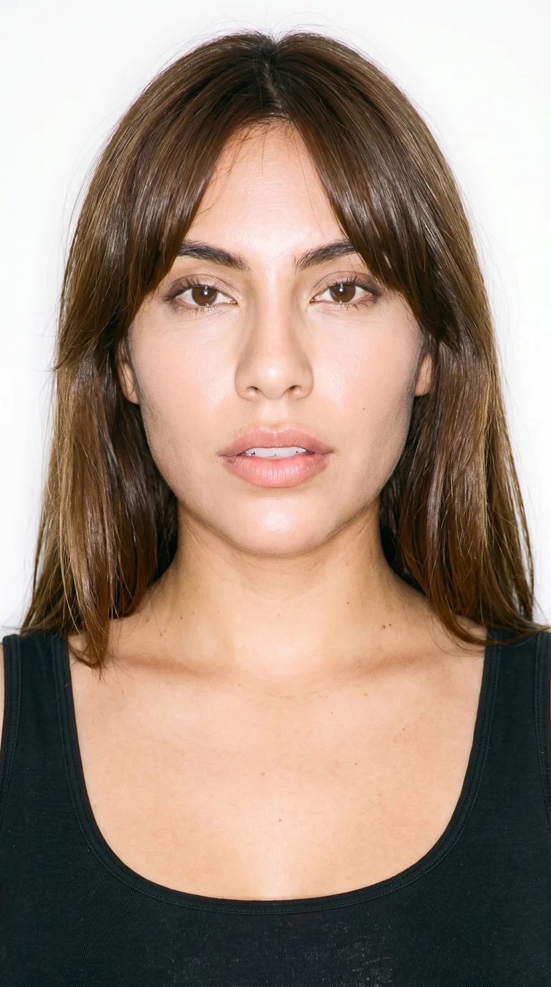 A 25-year-old Hispanic woman with medium olive skin and a neutral greenish-golden undertone. Her heart-shaped face has a wider forehead that narrows to a more pointed chin. She has thick eyebrows, brown eyes, and full lips. Her medium-brown hair is styled with longer curtain bangs parted in the center and framing both sides of her face.