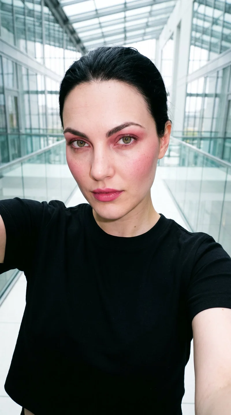 A 28-year-old Caucasian woman wears a black crop top in a front-facing selfie. Her head is tilted and she has a confident expression, with a skyscraper glass atrium backdrop.