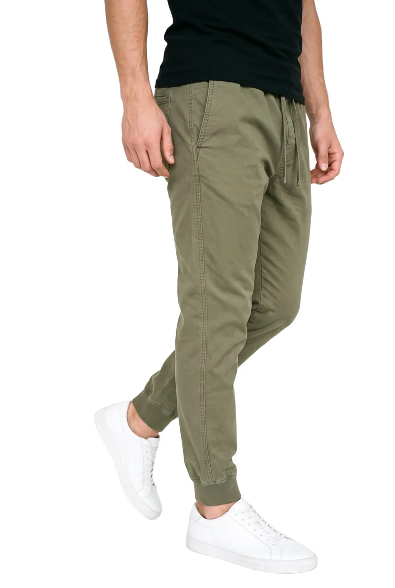 Olive men’s cuffed joggers shown on a male model from the lower body in an angled front view, with one heel lifted. The garment is presented against a pure white backdrop.
