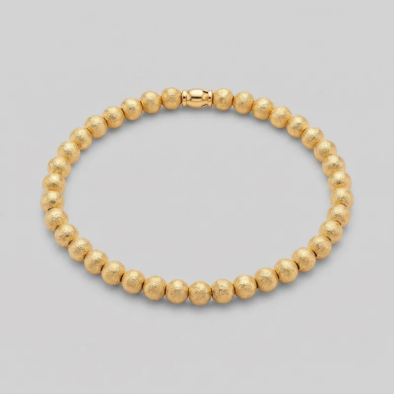 A complete gold beaded bracelet is arranged as an oval loop on a plain surface. It consists of closely spaced rounded beads with pebbled exterior surfaces and includes a smooth barrel-shaped closure element with narrow raised collars at each end.
