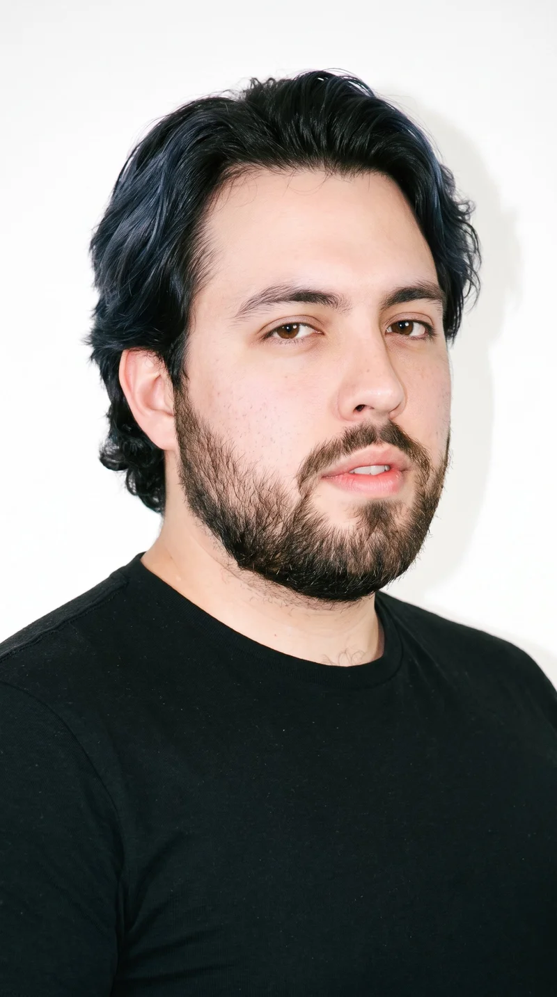A 25-year-old Caucasian man has lightly tanned fair skin with a peach undertone. His face is long, with a tall forehead and elongated cheeks, framed by thin eyebrows and brown eyes. He has medium lips and wavy, deep black hair with soft, relaxed movement. A short, full beard is evenly trimmed with clean cheek and neckline edges.