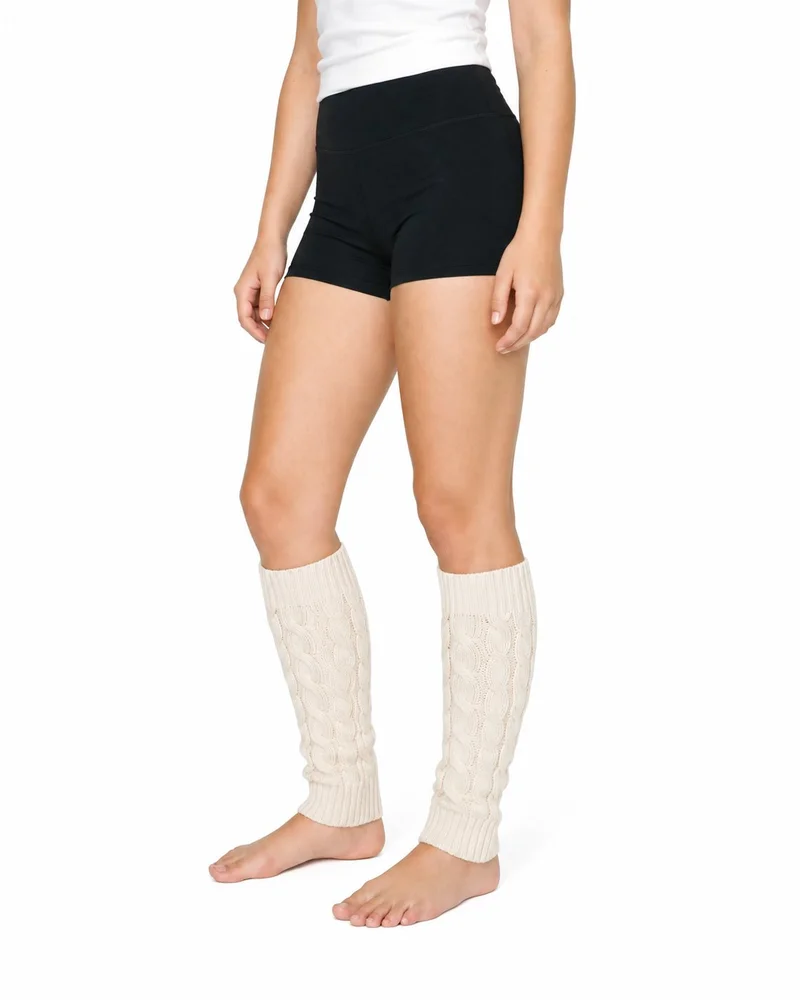 Cream cable-knit leg warmers shown on a female model’s lower legs from an angled front view, with her feet planted. The product is framed against a pure white backdrop.