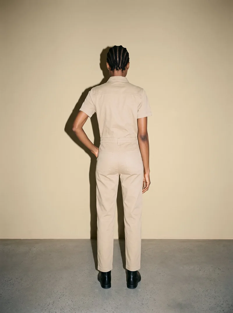 A 26-year-old African woman wears a khaki utility jumpsuit in a full-body rear view, standing with one hand on her hip. A sand dune backdrop fills the studio setting.