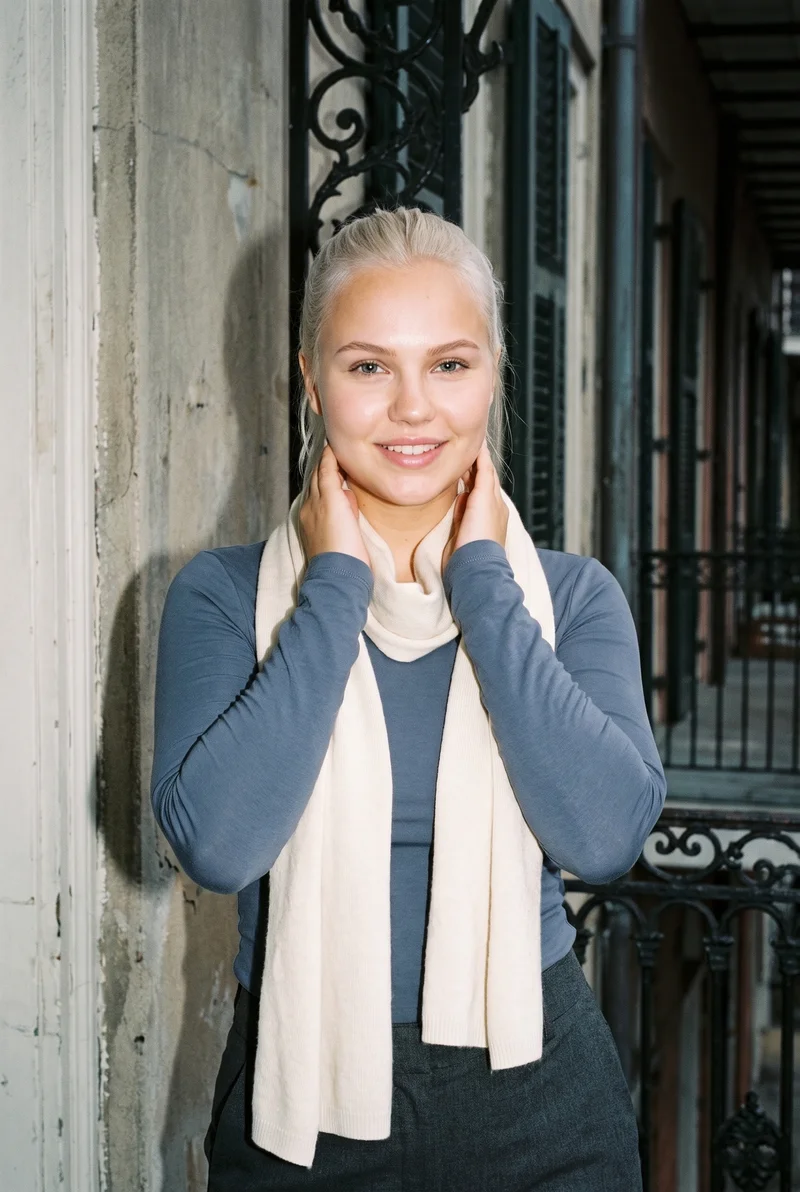 A 21-year-old Caucasian woman wears a cream knit wrap in an upper-body front view. She is smiling with her hands on her neck, against a New Orleans balcony backdrop.