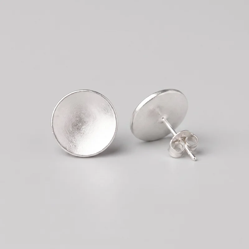 A pair of round silver disc stud earrings is shown as a standalone product. One earring faces forward, revealing its shallow domed circular face and raised rounded rim, while the other is angled to show its straight post and butterfly clutch backing. The disc surfaces have a subtly hammered, uneven reflective finish with no stones, logos, hanging elements, or additional embellishments.