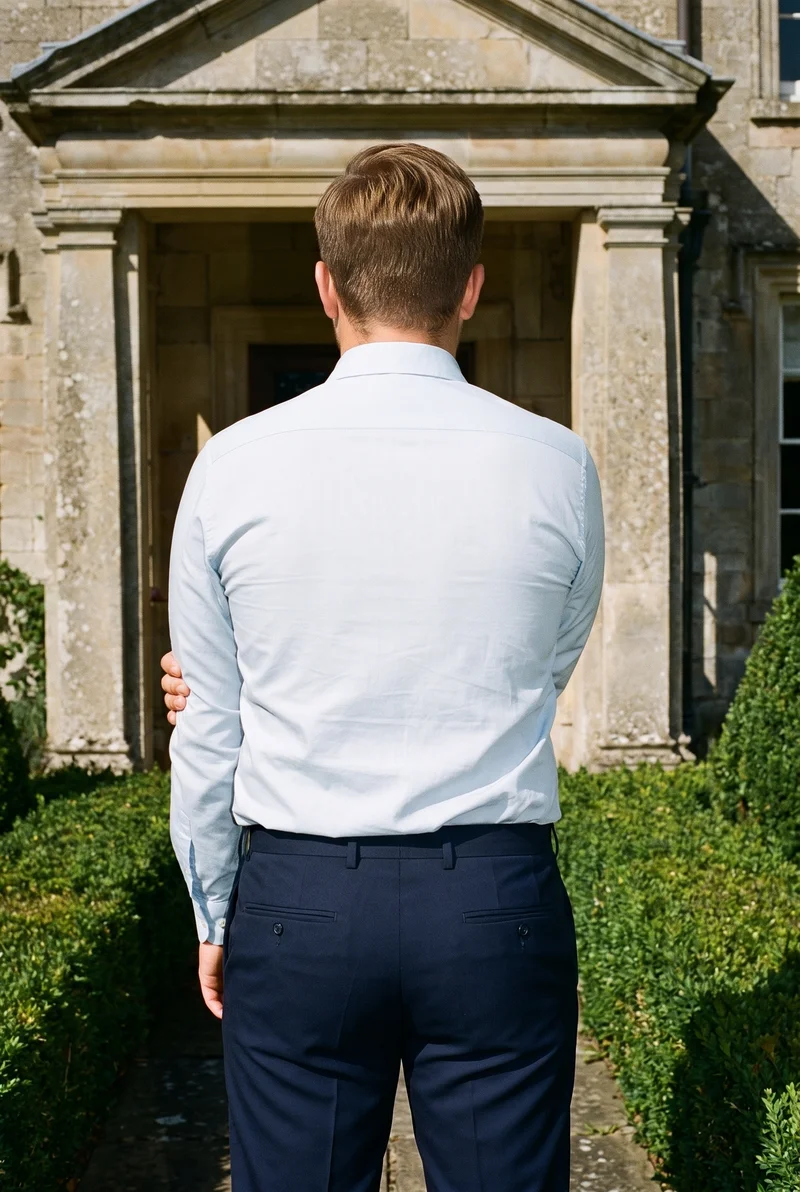 A 19-year-old Caucasian male model wears navy wool suit trousers in a three-quarter-length back view, holding one arm. An English countryside manor serves as the backdrop.