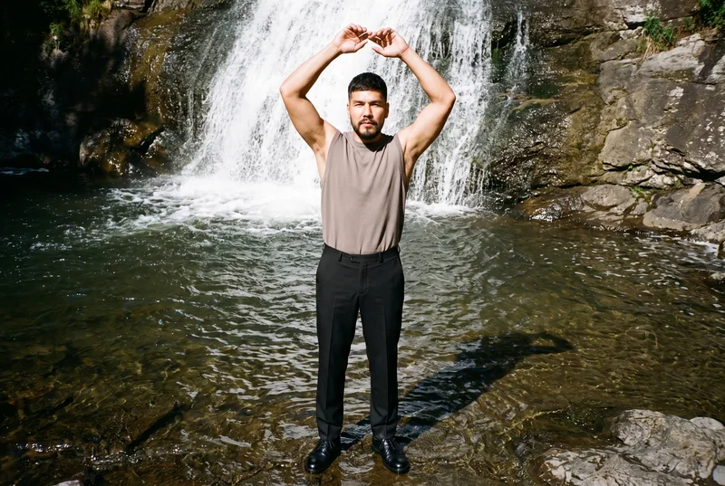 A 38-year-old Indigenous American man wears black tailored trousers in a front-view environmental composition. He stands with his arms overhead and a confident expression against a waterfall pool backdrop.