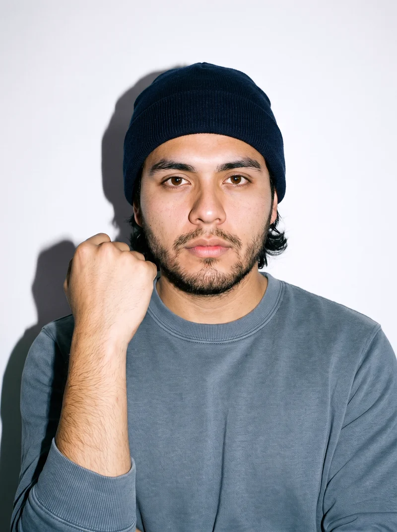 A 21-year-old Hispanic man is shown in a front-facing bust view wearing a navy beanie. His wrist is positioned at his jaw, and he has a serious expression. A porcelain studio-wall backdrop fills the background.