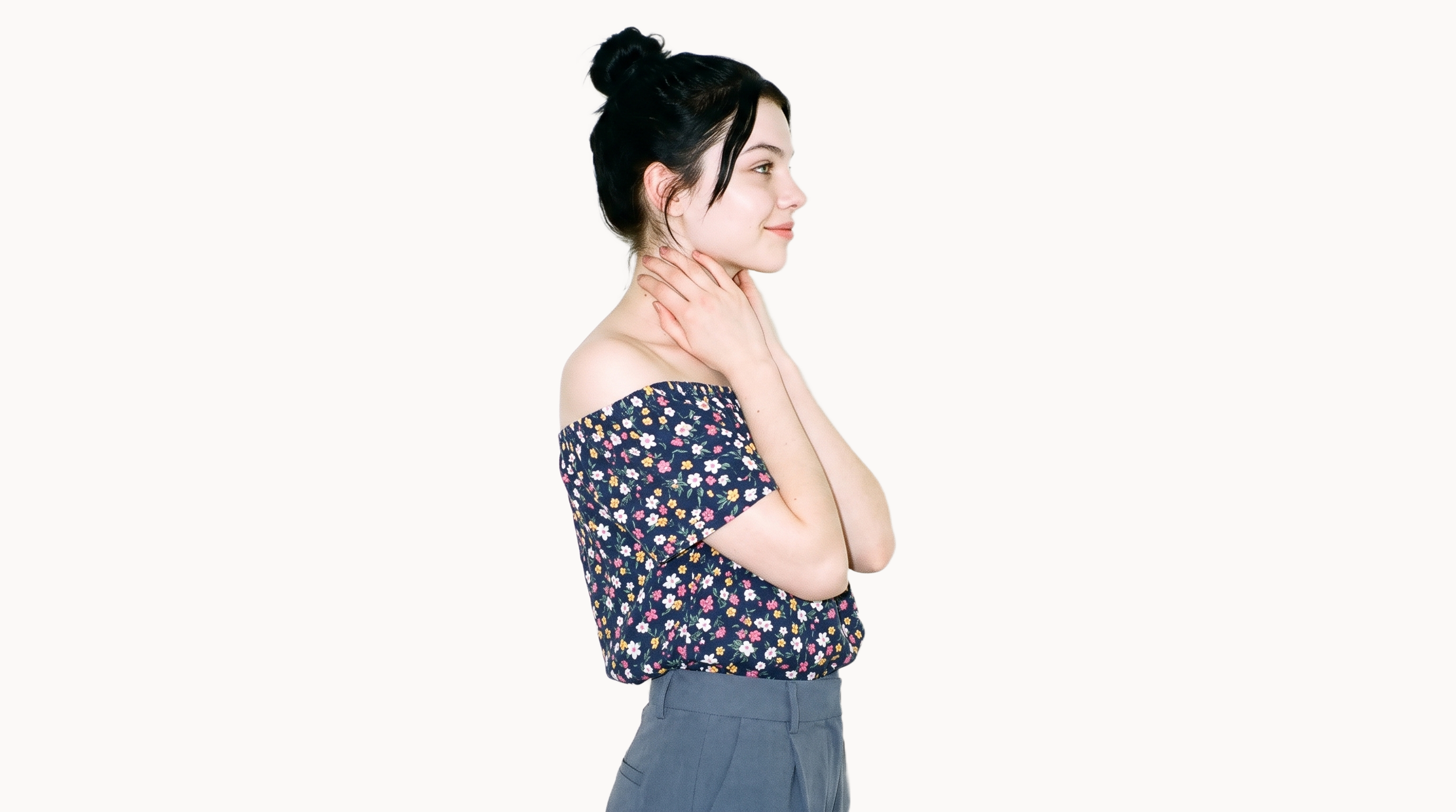 A 19-year-old Caucasian woman wears a floral print Bardot top in an upper-body side-view composition. She is smiling with her hands on her neck, against a flat Chantilly Lace backdrop.