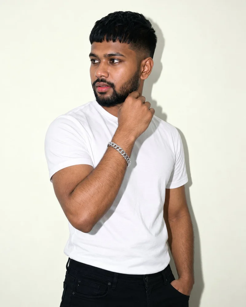 A 20-year-old Indian male model is shown in an angled upper-body view with his wrist held near his jaw, displaying a men's Cuban link bracelet. An ivory lace studio-wall backdrop frames the composition.