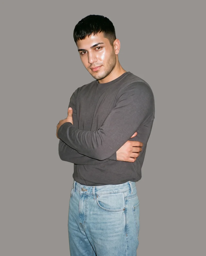 A 20-year-old Turkish male model wears men's light wash straight jeans in a three-quarter-length frame, shown from a front three-quarter angle in a self-hug pose with a natural expression. A flat grey studio backdrop surrounds the model.