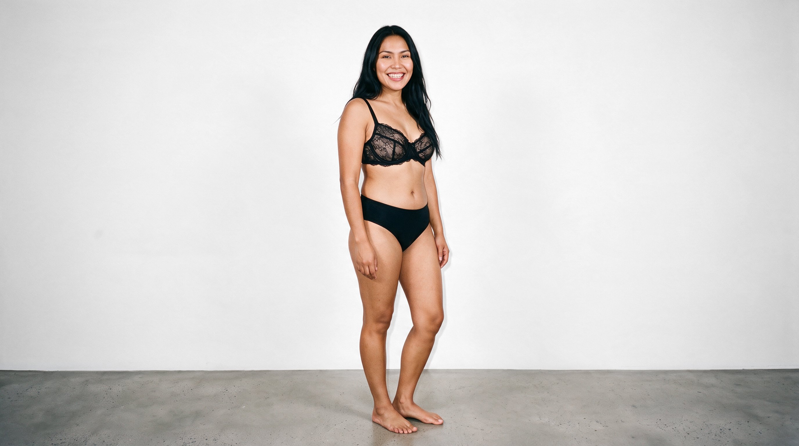 A 22-year-old Indigenous American woman wearing a black lace bra stands straight in a full-body, angled-front view. She has a grin, with a pure white studio-wall backdrop.