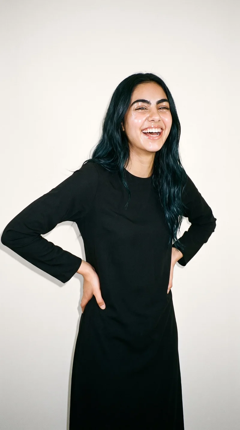 An 18-year-old Arab woman wears a black classic abaya against a soft ivory studio-wall backdrop. She is shown in an angled front view at three-quarter length, with her hands on her hips and a laughing expression.