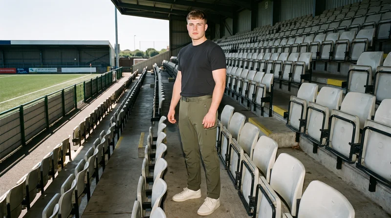 A 19-year-old Caucasian male model wears olive softshell hiking trousers in an environmental front three-quarter view, standing with his ankles crossed and a soft facial expression. Stadium seating forms the backdrop.