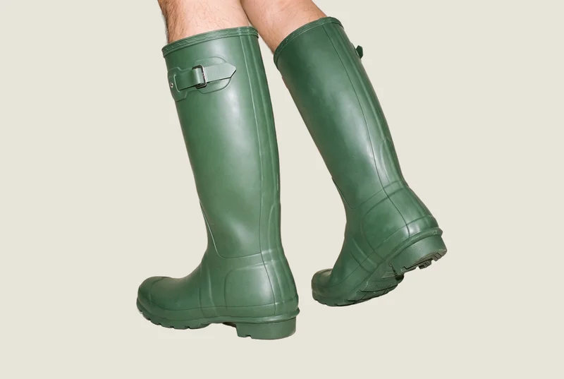 A pair of men's green Wellington boots shown from an angled rear view, with one foot positioned forward. Both feet are framed against a flat Oyster Pearl backdrop.