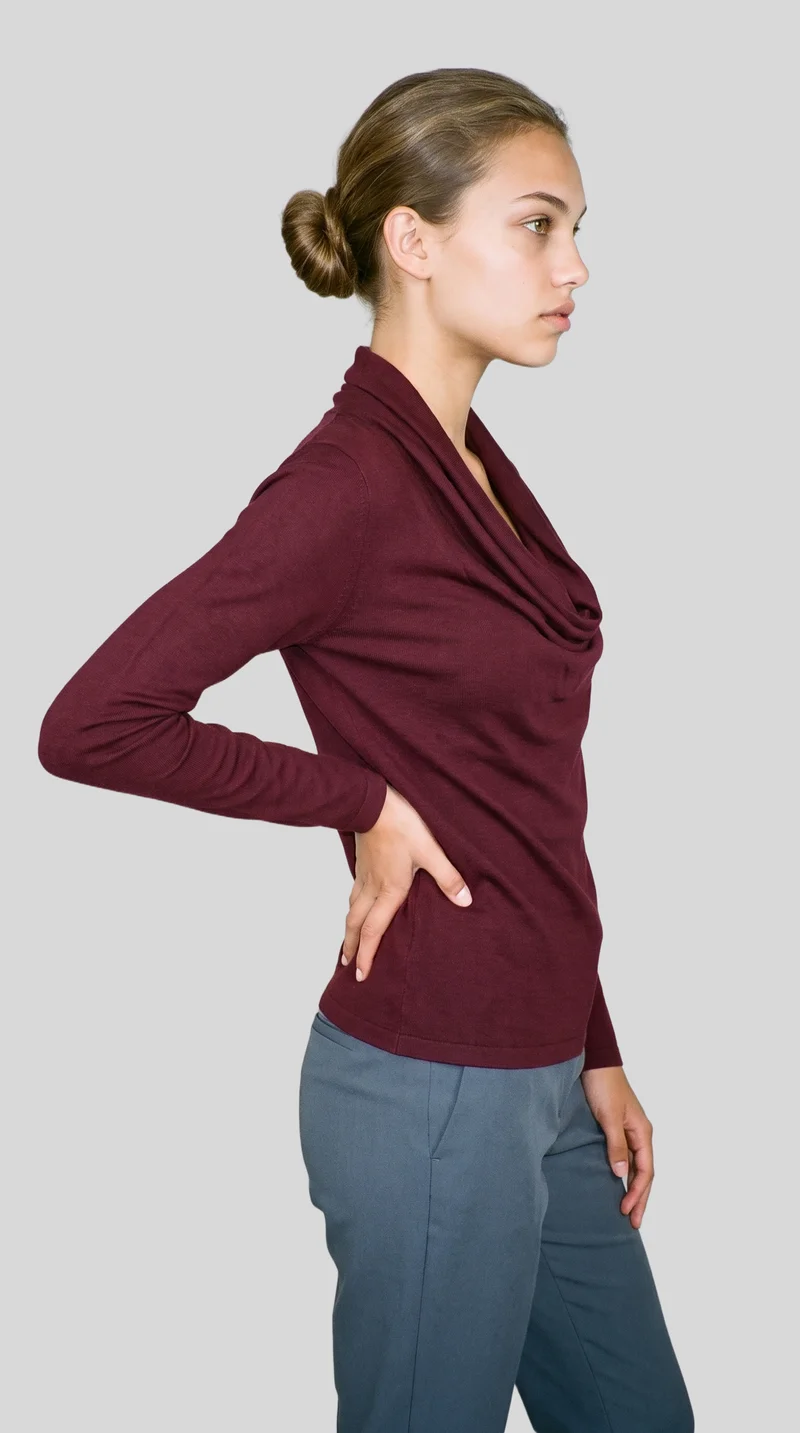 A 19-year-old Caucasian woman wears a burgundy cowl-neck top in a three-quarter-length side view, with one hand resting on her lower back and a neutral facial expression. The subject is shown against a flat pearl grey backdrop.