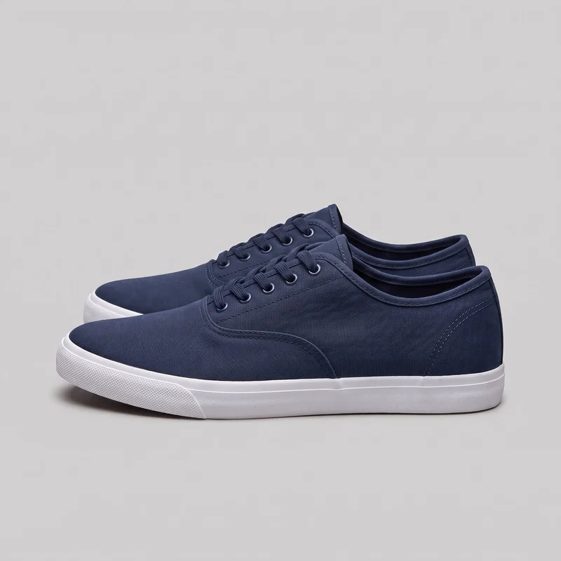 A pair of women's navy canvas sneakers is shown from an angled side/front view. The low-top shoes have rounded closed toes, plain lace-up uppers with stitched side panels, padded collars, flat soles with raised sidewalls, and textured toe bumpers.