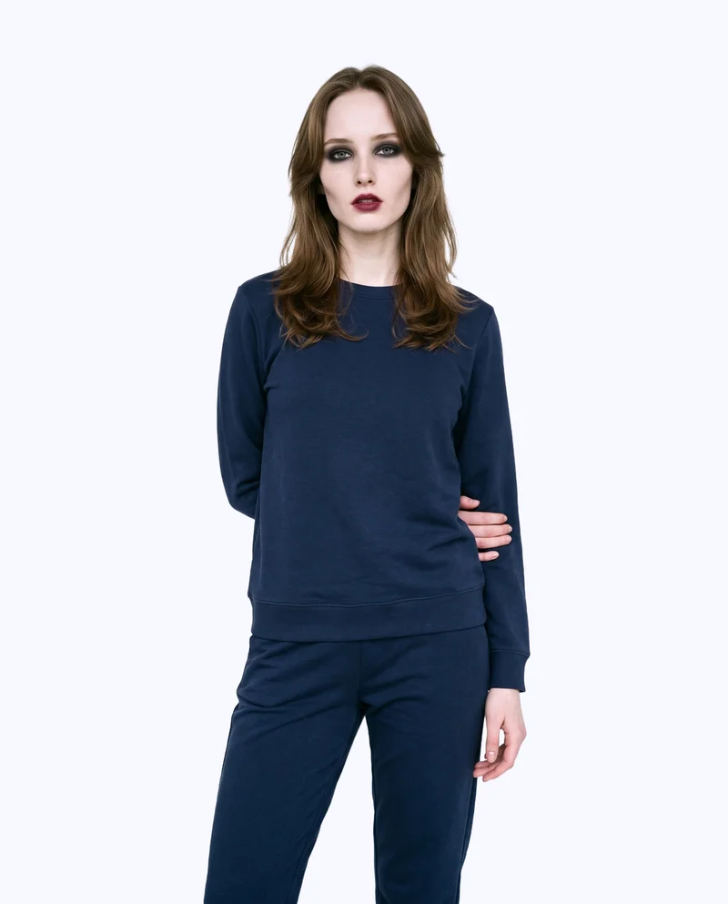 A 19-year-old Slavic woman wears a navy loungewear set in a three-quarter-length front view. One elbow is positioned behind her back, and she has a natural facial expression. The image uses a flat porcelain-colored backdrop.