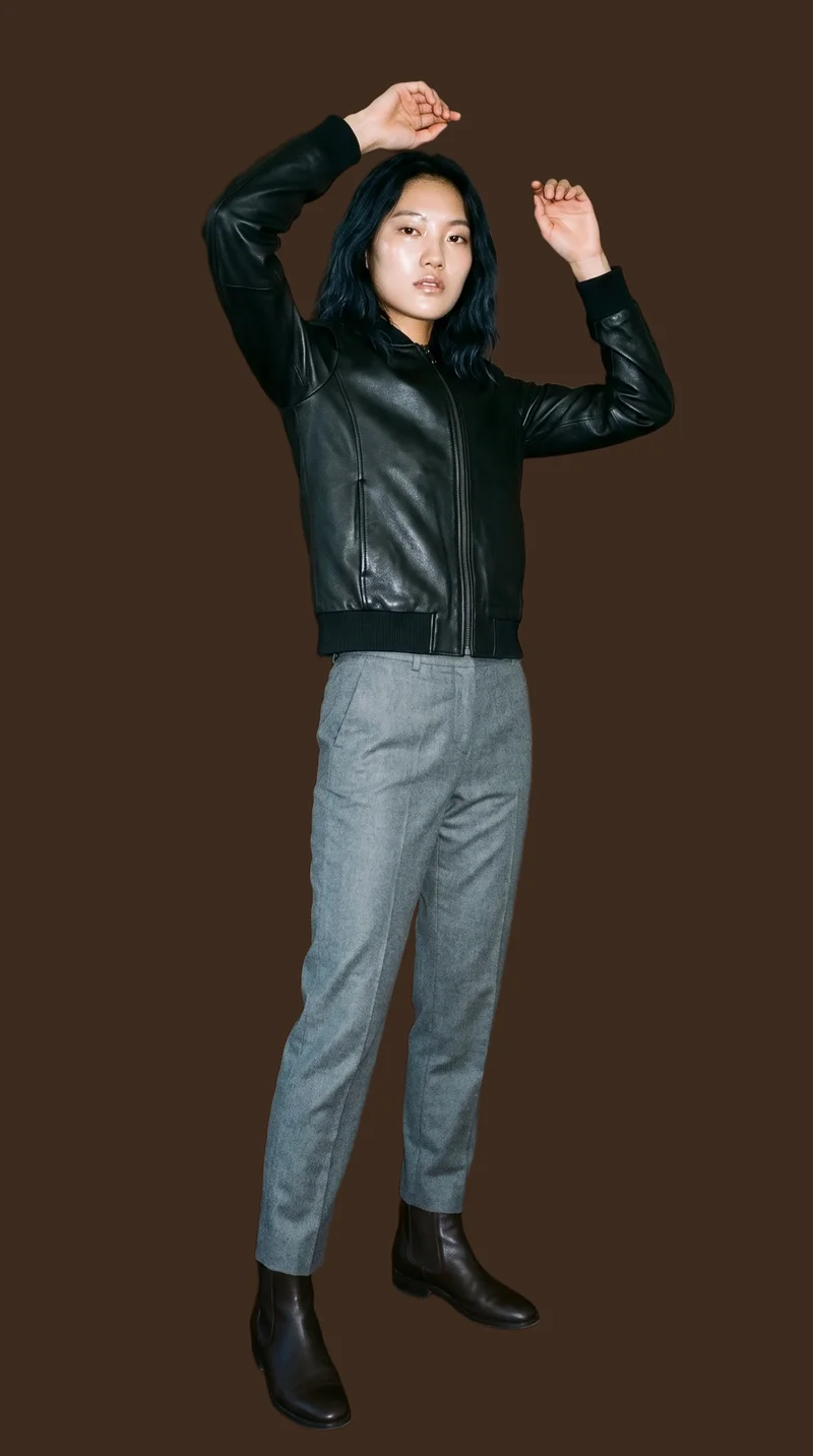 A 24-year-old Chinese woman wears a black bomber leather jacket in a full-body view from a front three-quarter angle. Her arms are raised overhead and her facial expression is neutral. An espresso flat-color backdrop frames the studio-cutout composition.