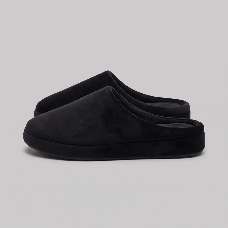 A pair of women's black memory-foam slippers is shown from a side three-quarter angle, with one closed-toe mule positioned in front of the other. The rounded toes, open heels, raised padded collar rims, mostly seamless uppers, and thick flat platform soles with rounded sidewalls are visible.
