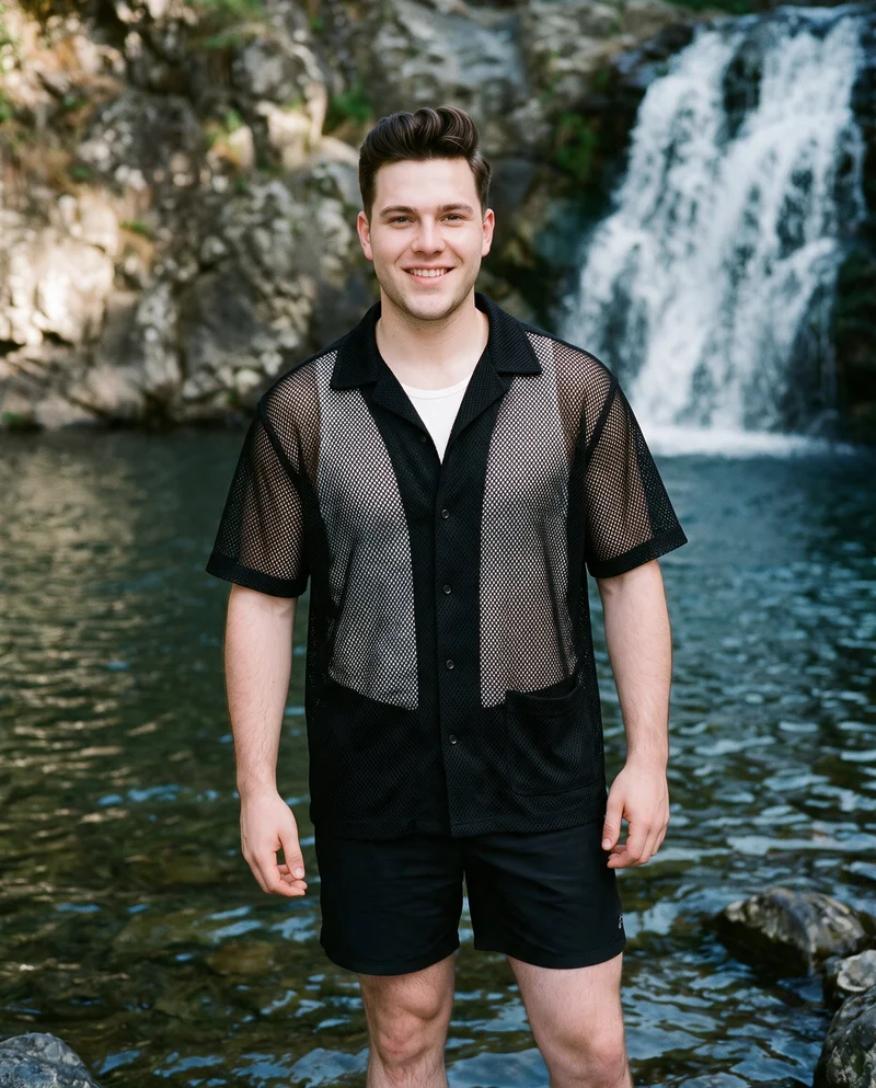 A 23-year-old Caucasian male model stands straight in a front-facing three-quarter-length view wearing a men's black mesh cover-up. He has a natural facial expression, with a waterfall pool backdrop.