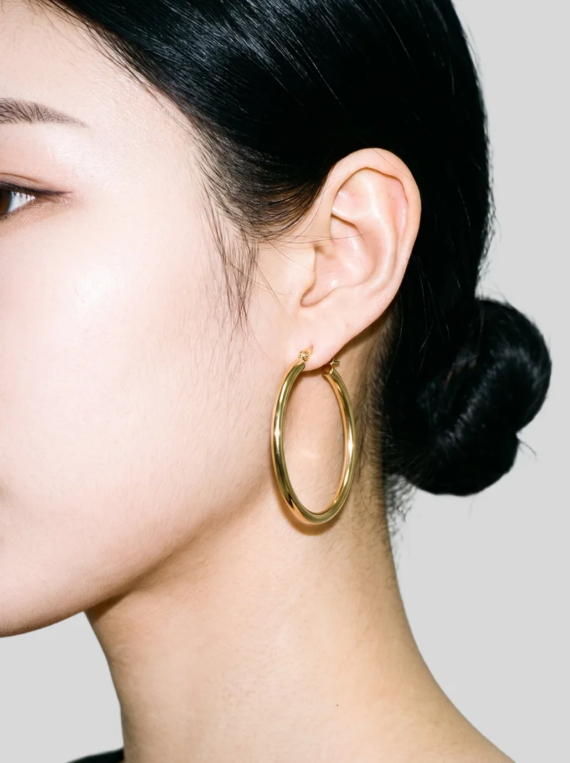 A gold hoop earring shown on an ear in side view, with the head held level. The image uses a flat pearl grey backdrop.