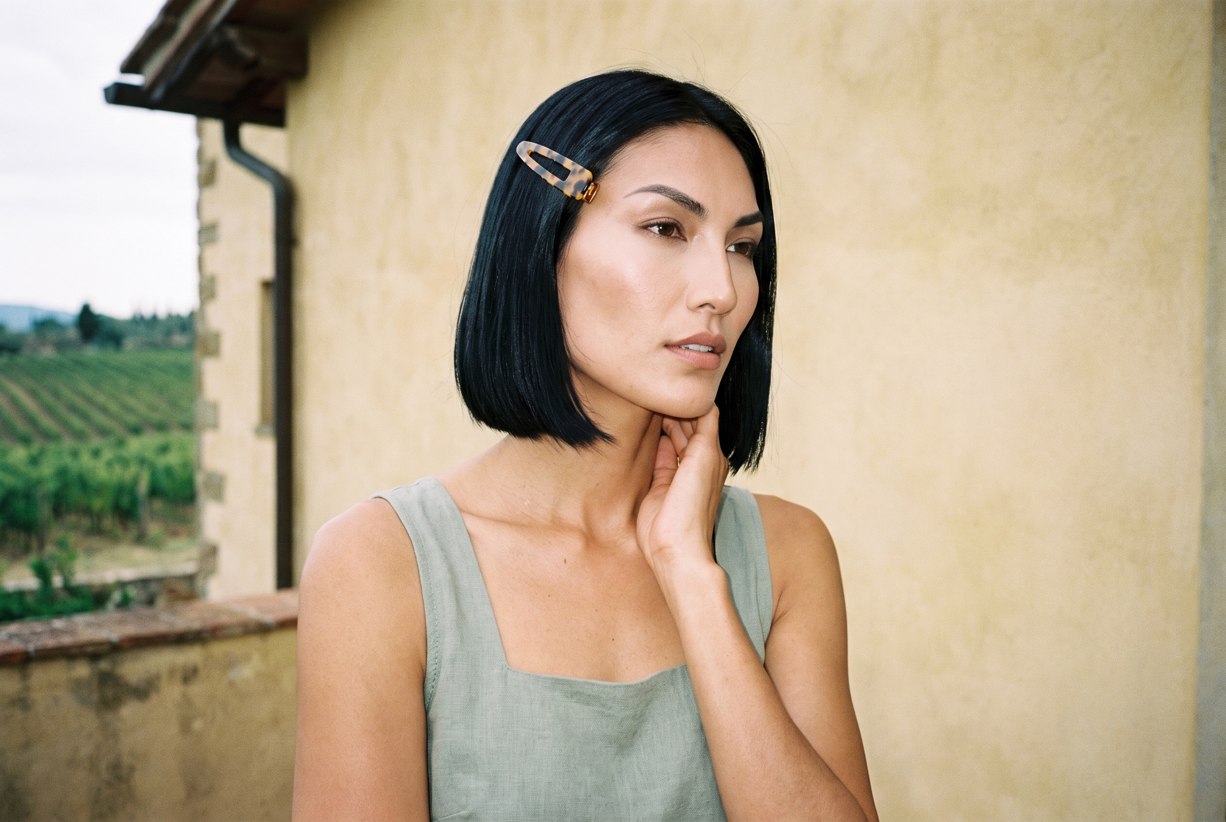 A 24-year-old Indigenous American woman is shown from the front in a bust-length view, wearing a tortoiseshell hair clip. Her hand rests on her neck, and she has a neutral expression. An Italian villa terrace serves as the backdrop.