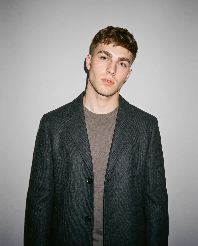 A 20-year-old Caucasian male model wears a charcoal overcoat in a front-facing bust composition with a shoulder-drop pose and neutral expression. A dove grey studio-wall backdrop frames the coat.