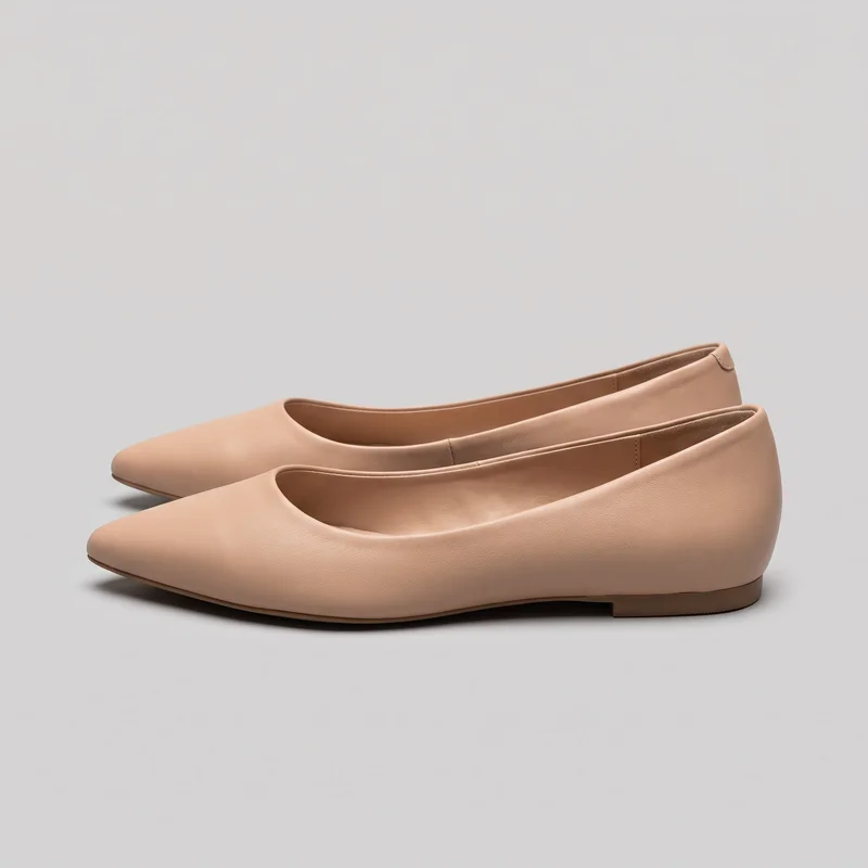 A pair of nude slip-on ballet flats is shown in a side three-quarter view, with both shoes mostly visible. The flats have tapered pointed toes, low-cut openings, smooth uppers, thin sole edges, and low stacked heels, with narrow piping around the openings and no visible closures or embellishments.