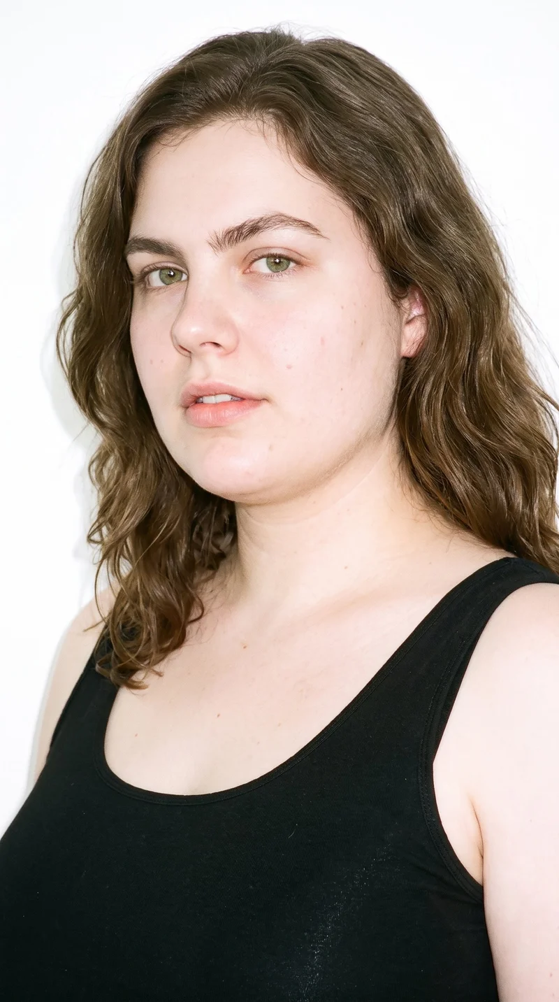 A 29-year-old Caucasian woman with pale skin and a neutral undertone has a soft, youthful baby face with full cheeks and rounded contours. She has thick eyebrows, brown eyes, and medium-sized lips. Her brown hair is styled in soft waves with gentle movement.