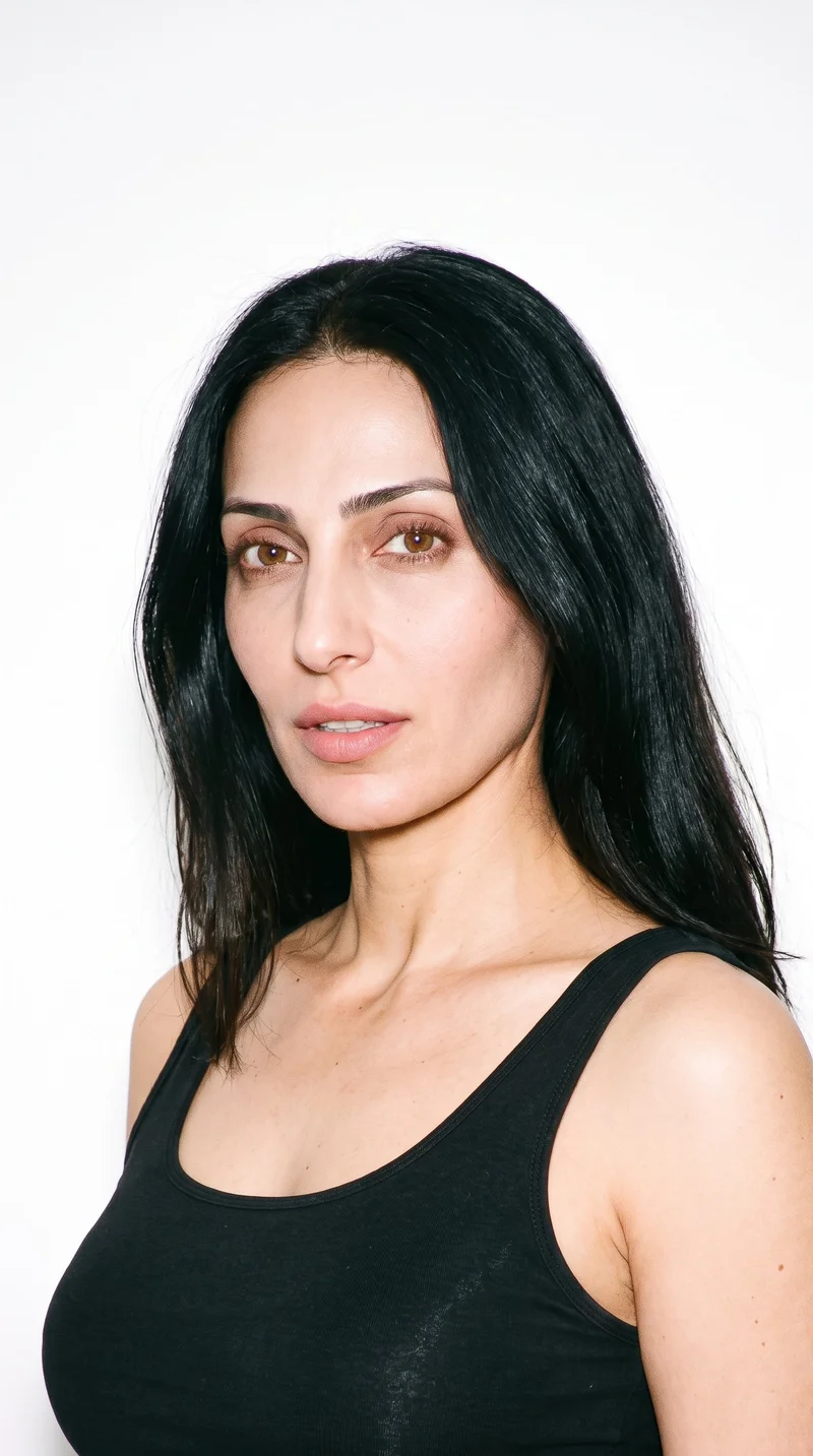 A 42-year-old Arab woman with honey-toned skin and a warm golden undertone. Her face has an inverted-triangle shape, with a wider forehead that narrows toward the chin. She has thick eyebrows, brown eyes, and full lips. Her long, deeply black hair is worn loose past the shoulders with natural volume.