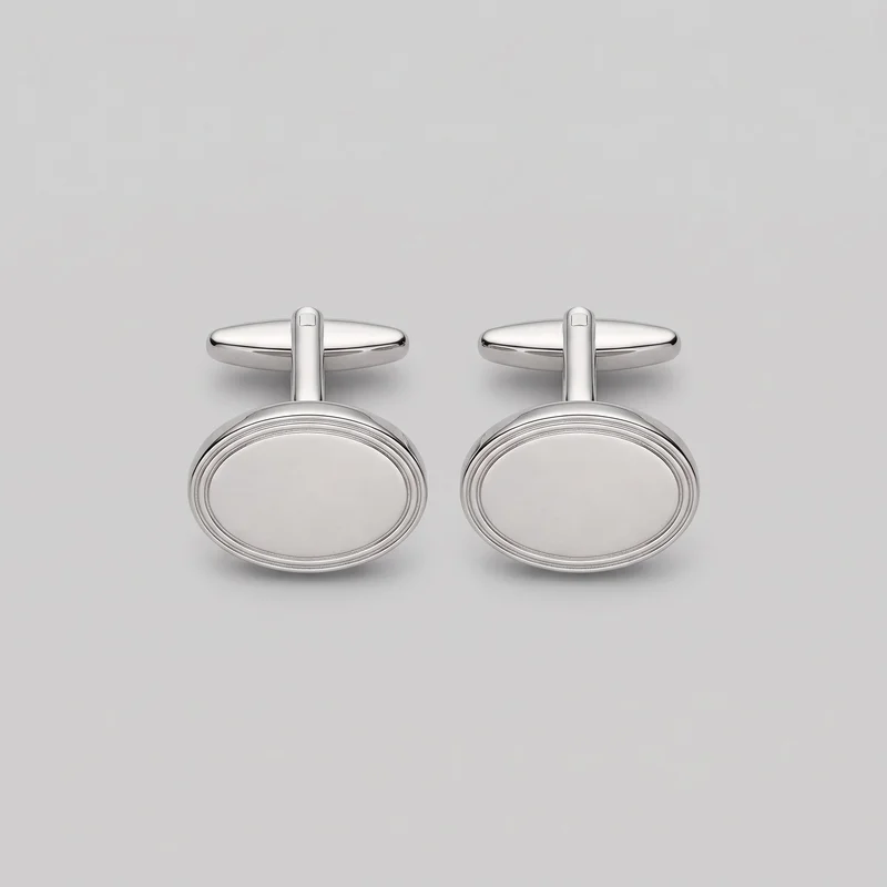 A matching pair of silver oval cufflinks stands upright on a plain surface. Each cufflink has a smooth domed oval front framed by multiple concentric raised rim lines, connected by a short vertical stem to a horizontal torpedo-shaped toggle back with rounded ends and a small rectangular inset on top.