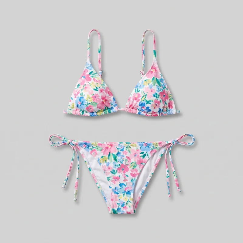 A two-piece string bikini set is arranged in a flat lay, with the triangle top positioned above the tie-side bikini bottom. The top has soft triangular cups with gathered lower edges, a narrow underbust string, slim adjustable shoulder straps, and a center front connection. The low-rise bottom has a triangular front panel, high-cut leg openings, narrow edge binding, and self-tie side strings knotted at both hips with trailing ends. An all-over floral and leaf print covers both pieces.