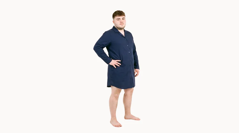 A 20-year-old Caucasian male model wears a navy cotton nightshirt against a Chantilly Lace flat-color backdrop. He is shown in an environmental frame from a front three-quarter angle with one hand on his hip and a confident expression.