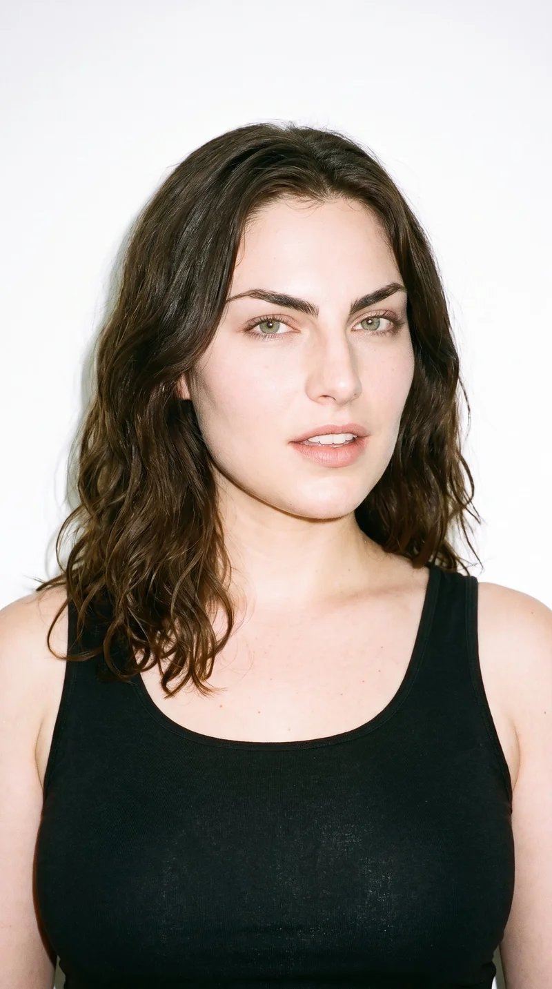 A 23-year-old Caucasian woman with very pale skin and a peach undertone. Her diamond-shaped face has a narrow forehead and jaw and is widest at the cheekbones. She has medium-thickness eyebrows, green eyes, and thin lips. Her dark-brown hair is styled in soft, natural waves.