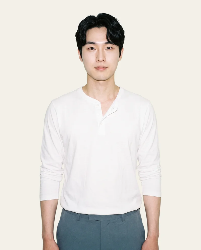 A 27-year-old Korean man wears a white Henley top in an upper-body, front-facing view with his arms relaxed and a confident expression. The image uses an alabaster flat-color backdrop.