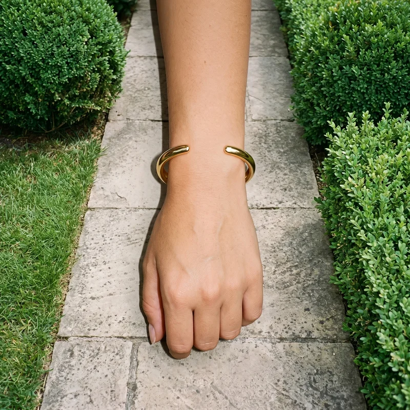 A woman's hand and wrist are shown from above with the knuckles facing forward, wearing a gold cuff bracelet. A topiary garden serves as the backdrop.