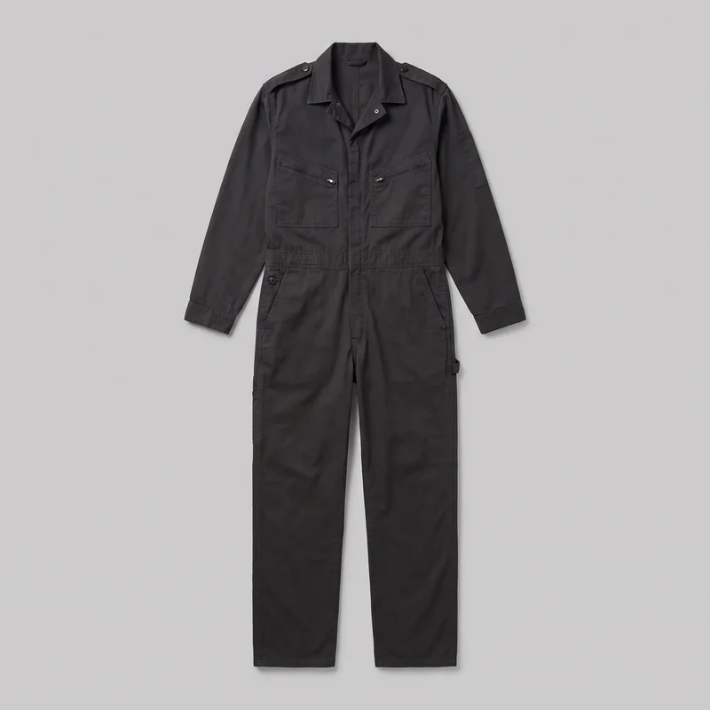 A black men's utility coverall jumpsuit is shown in a full standalone front flat-lay view. It has a pointed spread collar, long sleeves with straight cuffs, shoulder epaulettes, a concealed front closure, a stitched waistband seam, straight full-length legs, two chest patch pockets with diagonal zip openings, front hip pockets, a sleeve patch pocket, and a leg utility tab. Prominent topstitched panel seams are visible throughout.