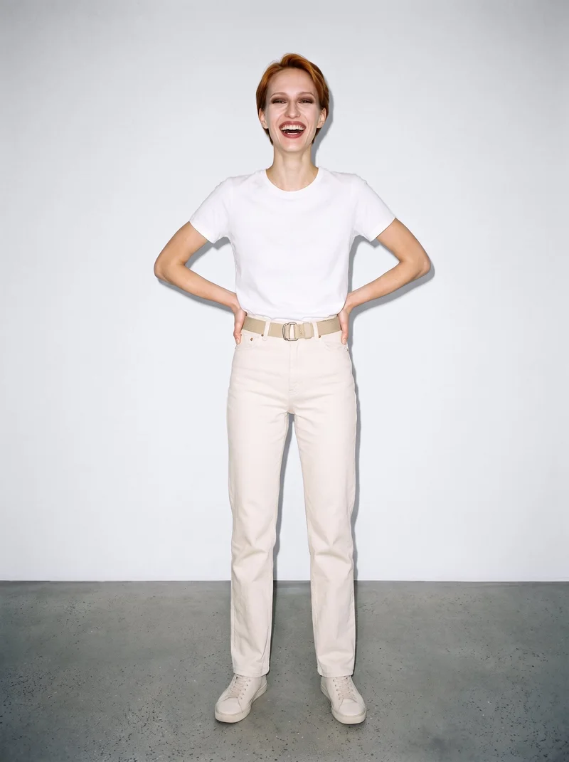 A 21-year-old Caucasian woman is shown full body from the front, laughing with her hands on her hips while wearing a beige canvas belt. A porcelain studio-wall backdrop fills the background.