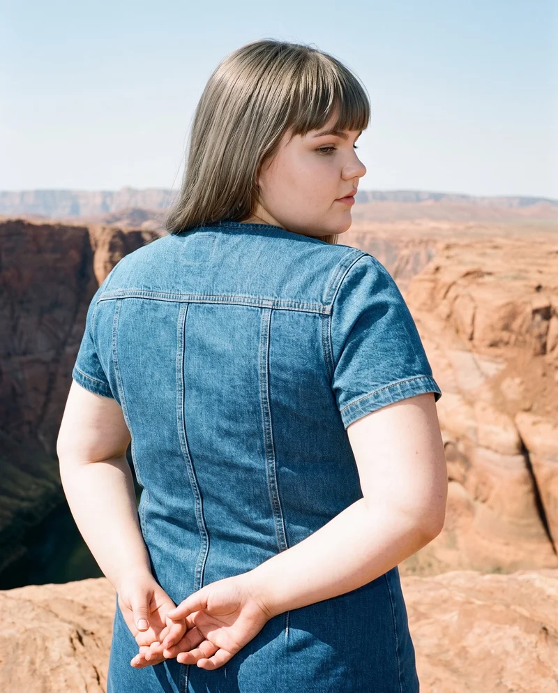 A 20-year-old Caucasian woman wears a denim mini dress, shown from the upper body in an angled rear view with her hands behind her back. The Grand Canyon Rim appears as the backdrop.