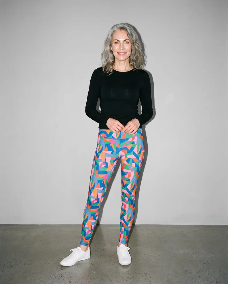A 55-year-old Caucasian woman models printed leggings in a full-body front view, standing with her hands in front and a natural facial expression. A pebble grey studio-wall backdrop fills the background.