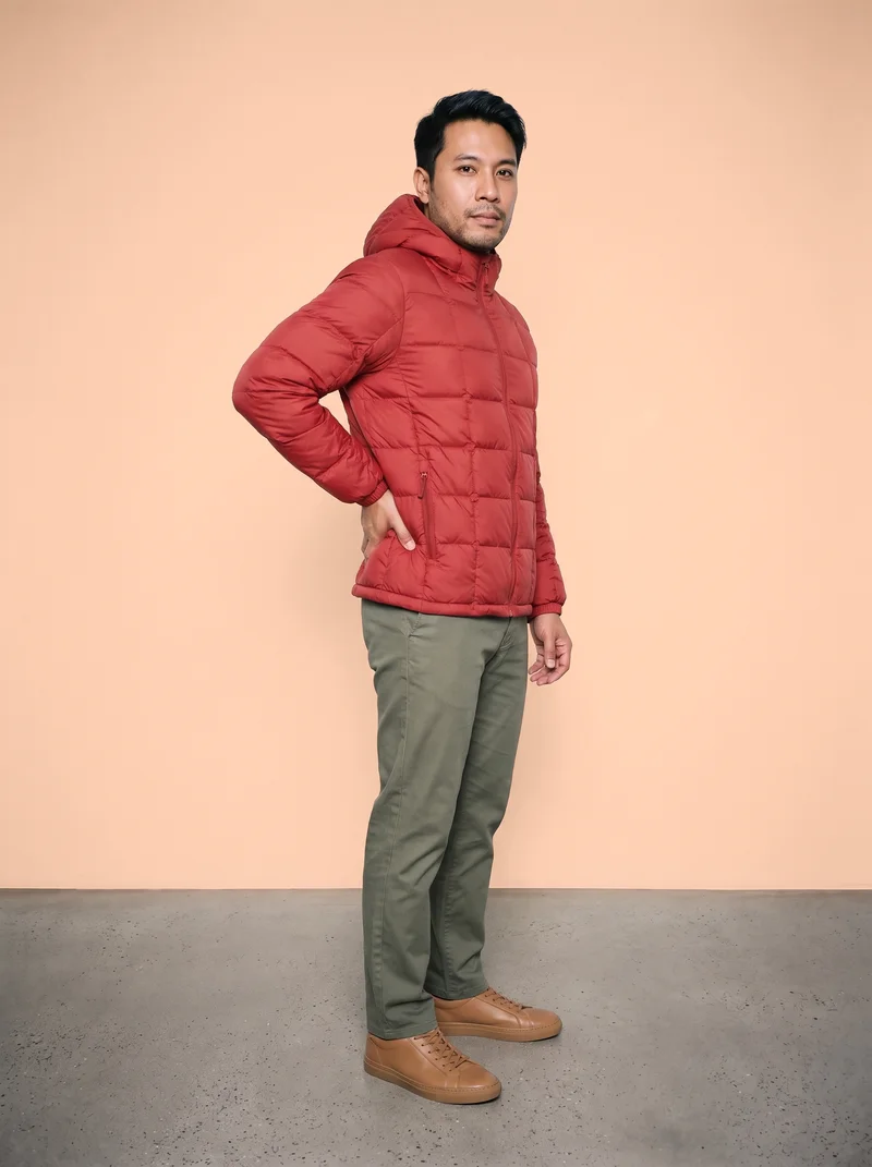 A 42-year-old Southeast Asian man wears a red quilted down jacket in a full-body front three-quarter view. He stands with one hand on his lower back and a neutral facial expression against an apricot studio-wall backdrop.