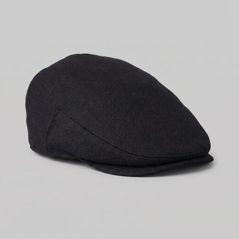 A mostly complete black flat cap is shown alone in a three-quarter view. It has a low-profile rounded crown, curved side panel seams, a shaped side section, a front seam following the brim line, and a short forward visor tucked beneath the front edge. The cap has a subtly raised crown ridge and a fine woven surface texture, with no visible logos or embellishments.