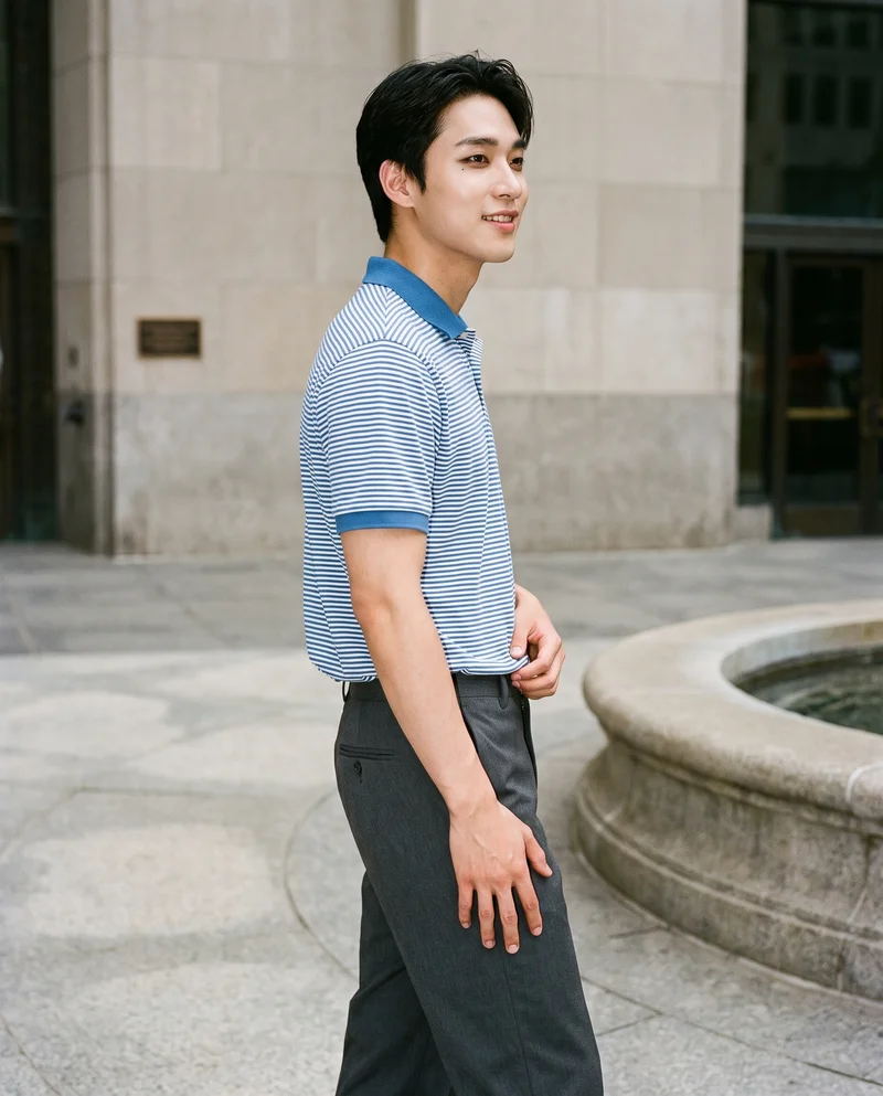 A 21-year-old Korean man wears a striped cotton polo shirt in a three-quarter-length side view, posed with one hip out and smiling. A Fountain Plaza backdrop is visible behind him.