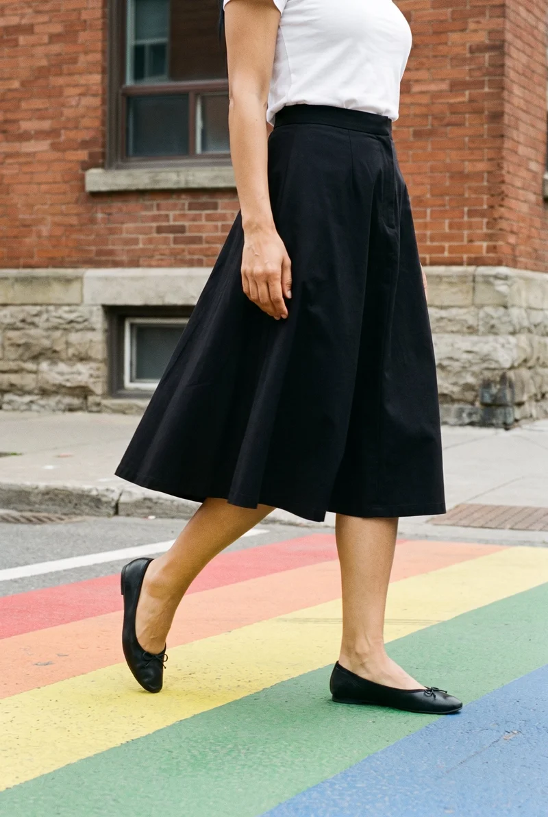 A black A-line skirt shown on a female model from an angled front view, framed on the lower body as she walks. A rainbow crosswalk and Victorian houses form the backdrop.