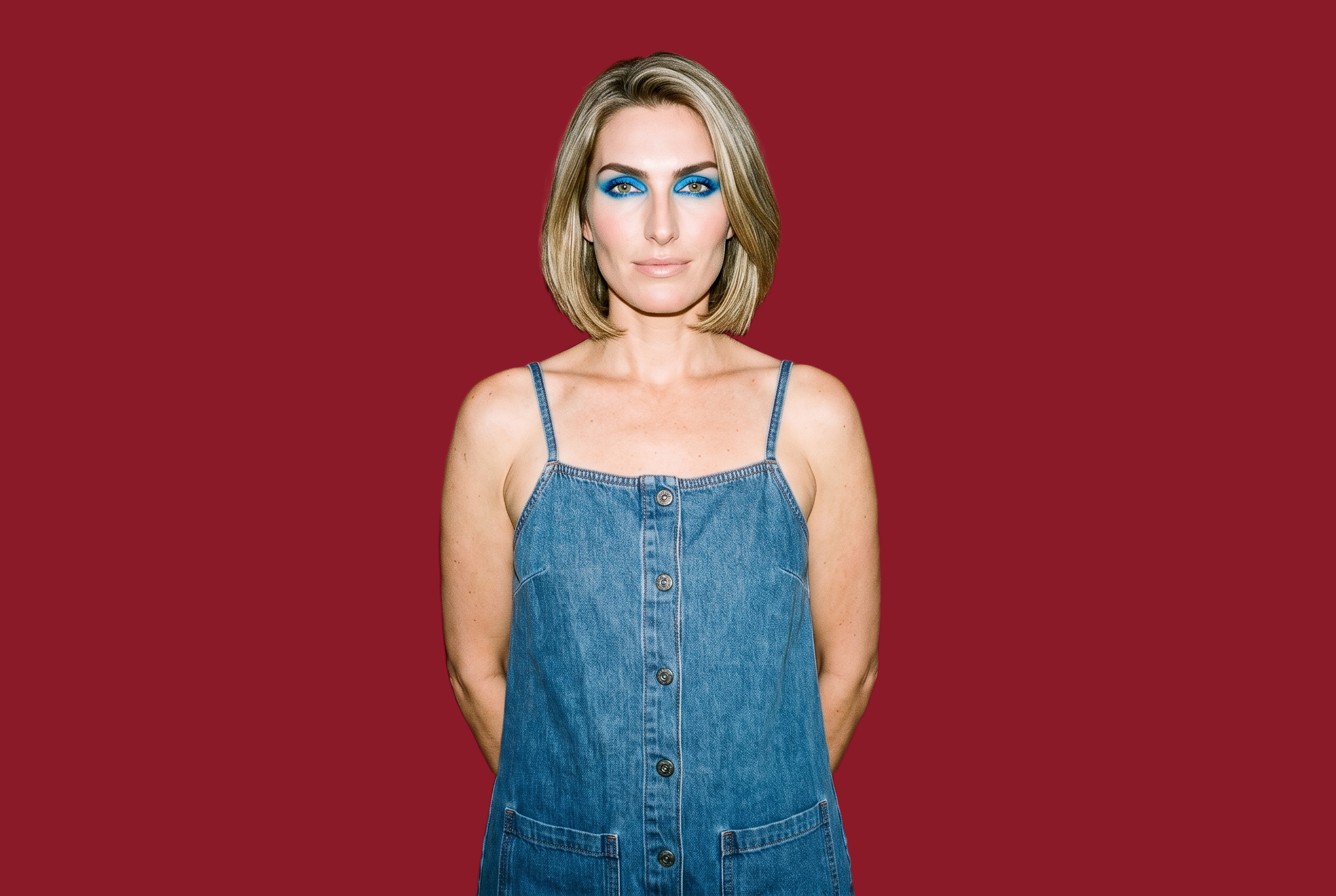 A smiling 40-year-old Caucasian woman wears a denim playsuit in an upper-body, front-facing view, with her hands behind her back. A flat crimson backdrop frames the studio-cutout composition.